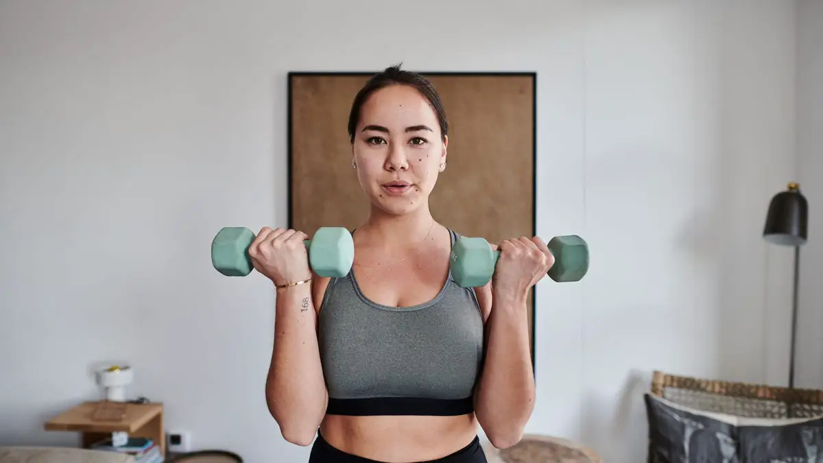 A person with pulled-back brown hair in a grey sports bra lifting light green hand weights.